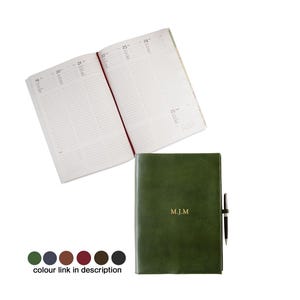 May include: An open, lined notebook with a red ribbon bookmark is positioned above a closed, dark green leather journal. The closed journal has the initials "M.J.M" in gold lettering and a pen attached. A color palette of green, blue, brown, red, and black is shown.
