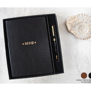 May include: A black rectangular notebook with the name BENJI embossed in gold lettering between two small stars, presented in a black gift box alongside a matching black and gold ballpoint pen. A fossilized ammonite shell sits nearby.