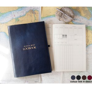 May include: A blue leather-bound logbook with gold lettering that reads "Klaus A. Wolf Logbook". The logbook is open to a blank page with a grid pattern. The logbook is on a table with a nautical chart and a pen.