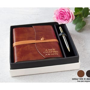 Könnte beinhalten: Geschenkset mit einem braunen Lederjournal mit dem Text "A New Chapter Awaits" und einem silbernen Stift, präsentiert in einer schwarz-weißen Box. Eine rosa Rose dient als Dekoration.