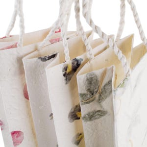 K&ouml;nnte beinhalten: Satz mit f&uuml;nf wei&szlig;en Papiert&uuml;ten mit Blumendesigns und wei&szlig;en Kordelgriffen. Die Taschen haben verschiedene Blumenmuster, darunter rosa Blumen, gr&uuml;ne Bl&auml;tter und gelbe Blumen.