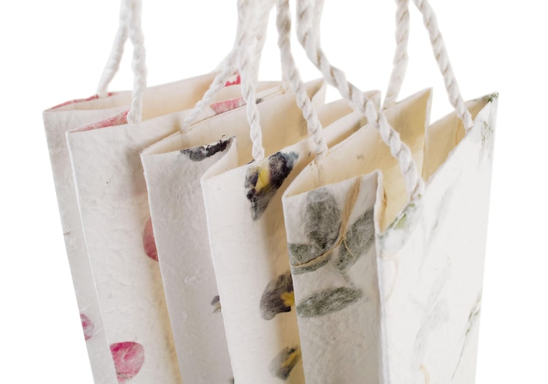 K&ouml;nnte beinhalten: Satz mit f&uuml;nf wei&szlig;en Papiert&uuml;ten mit Blumendesigns und wei&szlig;en Kordelgriffen. Die Taschen haben verschiedene Blumenmuster, darunter rosa Blumen, gr&uuml;ne Bl&auml;tter und gelbe Blumen.