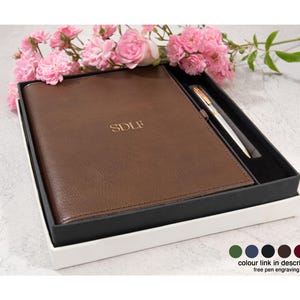 May include: A brown leather-textured notebook with the letters SDLF embossed in gold on the cover, presented in a black box alongside a matching metallic pen. The set is decorated with pink flowers and includes a color selection chart.