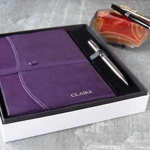 May include: A purple leather journal with the name "CLAIRE" embossed on the cover, a silver pen, and a red agate paperweight in a white gift box.