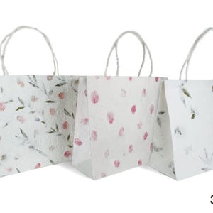 K&ouml;nnte beinhalten: Drei wei&szlig;e Papiert&uuml;ten mit Blumenmustern. Die Taschen haben wei&szlig;e Henkel und sind mit rosa und gr&uuml;nen Blumendesigns verziert.