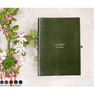 May include: A dark green leather journal with the text "J. DAWN VOLUME 1" in gold lettering. The journal is closed with a strap. The background features a sprig of a flowering tree with pink and white blossoms.
