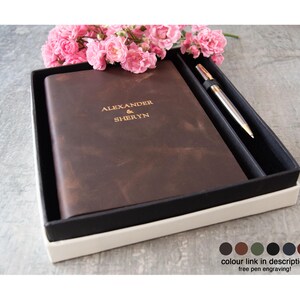 May include: A dark brown leather journal with the gold embossed text ALEXANDER & SHERYN, paired with a metallic pen in a black presentation box. The set is decorated with pink flowers and includes a colour selection chart at the bottom right.