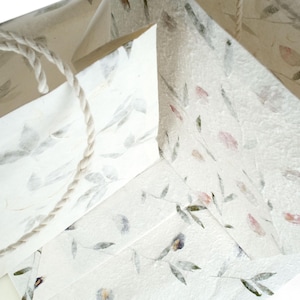 K&ouml;nnte beinhalten: Drei wei&szlig;e Papier-Geschenkbeutel mit Blumenmuster und einem Seilhenkel. Die Beutel sind aus recyceltem Papier mit eingebetteten Blumen und Bl&auml;ttern.