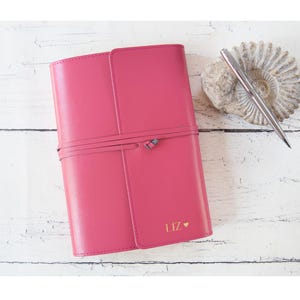 Pode incluir: Um diário de couro rosa choque com um fecho de cordão a condizer. O diário é personalizado com o nome "LIZ" e um símbolo de coração em dourado. Uma caneta prateada repousa sobre uma pedra decorativa ao lado do diário.