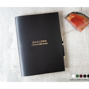 Puede incluir: Una carpeta negra de aspecto cuero con el texto JEAN LEWIS COACH PERSONNEL grabado en dorado en la portada. Un bolígrafo fino negro y dorado está insertado en el lateral de la carpeta y una rodaja de ágata pulida naranja y blanca está cerca.