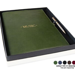May include: A dark olive green leather-bound journal with the word MUSIC embossed in gold on the cover, presented in a black box alongside a silver-toned metal pen. Below the box are six small colored circles and the text colour link in description.