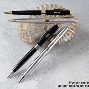 three pens with personalized names engraved on them, placed on a rock or seashell.