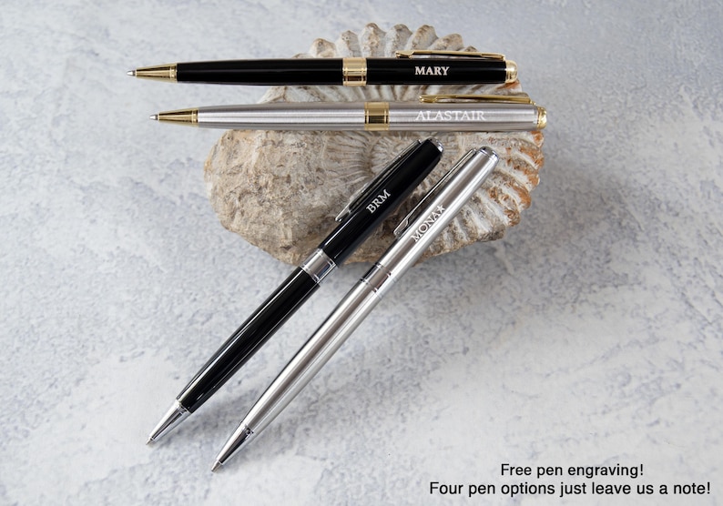 three pens with personalized names engraved on them, placed on a rock or seashell.
