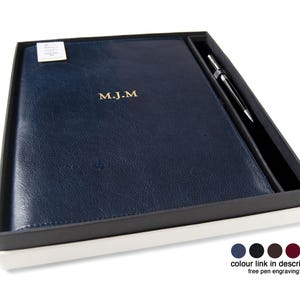 May include: A dark blue leather-like notebook with the gold embossed initials M.J.M. on the cover, presented in a rectangular box alongside a silver-toned metal pen. The set includes a small white tag and a color selection guide at the bottom.