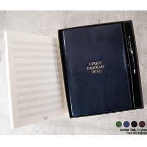 May include: A dark blue leather-bound notebook with the gold text LYRICS INSIDE MY HEAD on the cover, presented in a box with a matching pen. The open notebook displays cream-colored pages with printed musical staves for writing music.
