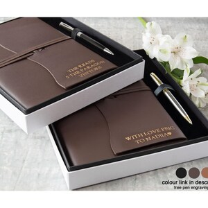 May include: Two brown leather journal and pen sets presented in white boxes. Each set includes a pen and a journal with text. One journal reads "THE READS 5 THE PARAGON VISITORS". The other reads "WITH LOVE PBIC TO NADIA".