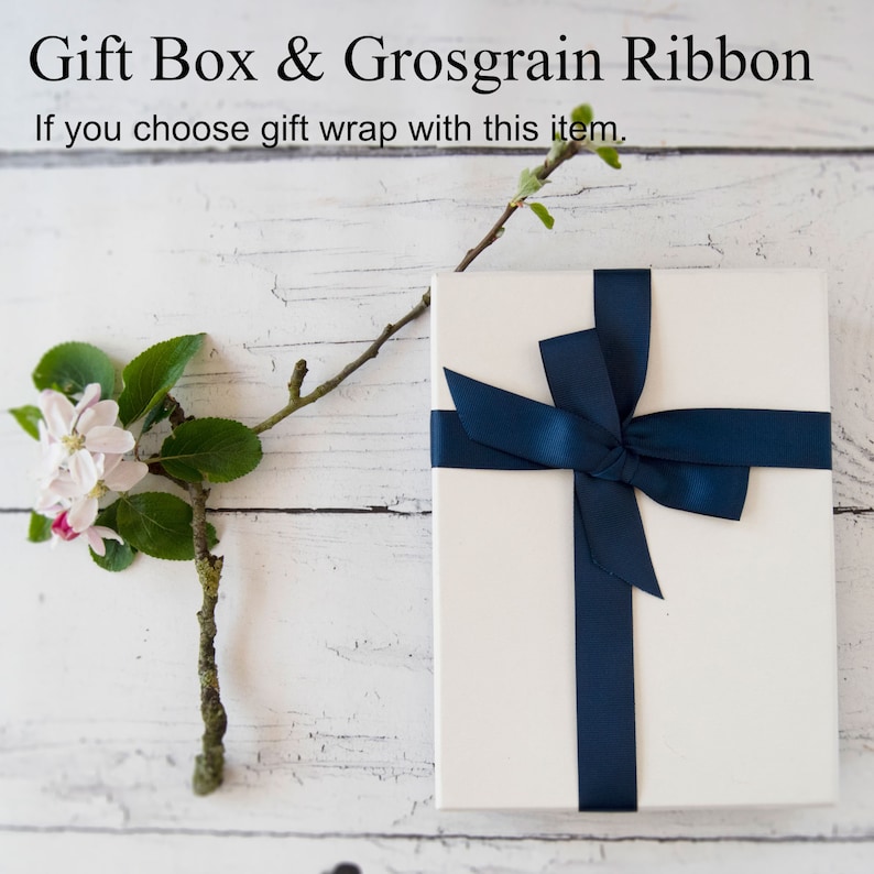 May include: A white gift box with a navy blue grosgrain ribbon tied in a bow. The box is sitting on a white wooden surface with a small branch of a flowering tree to the left.