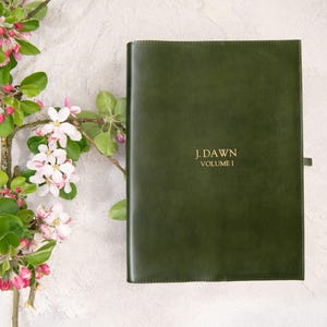 Puede incluir: Un libro encuadernado en cuero verde oscuro con letras doradas que dicen "J. Dawn Volume I". Una rama de un &aacute;rbol en flor con capullos rosas y flores blancas en el fondo. El libro tiene una cinta de marcador.