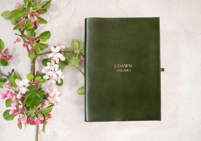 Puede incluir: Un libro encuadernado en cuero verde oscuro con letras doradas que dicen "J. Dawn Volume I". Una rama de un &aacute;rbol en flor con capullos rosas y flores blancas en el fondo. El libro tiene una cinta de marcador.