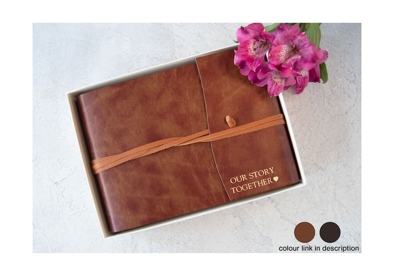 Puede incluir: Un diario encuadernado en cuero marr&oacute;n con una correa de cuero marr&oacute;n claro y las palabras "Our Story Together" grabadas en oro. El diario est&aacute; en una caja blanca y una flor rosa est&aacute; en el fondo.