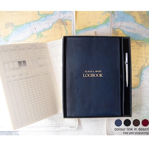 May include: A dark blue logbook with the gold text KLAUS A. WOLF LOGBOOK on the cover, presented in a black box with a silver pen. An open logbook page with printed tables and columns is visible to the left, showing fields for date, time, and notes.
