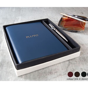 Op de afbeelding: Een donkerblauw notitieboek met het woord PLUTO in goud gedrukt op de kaft, gepresenteerd in een zwart-witte doos met een zilveren pen. Naast de doos liggen twee pennen op een gepolijste, gestreepte oranje en bruine agaatsteen presse-papier.