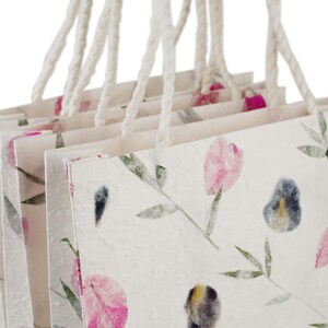 K&ouml;nnte beinhalten: Ein Stapel wei&szlig;er Geschenkt&uuml;ten mit Blumendesigns. Die Taschen haben wei&szlig;e Kordelgriffe und sind aus strukturiertem Papier gefertigt. Die Blumendesigns sind rosa, gr&uuml;n und blau.