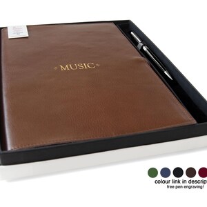 May include: A brown leather-textured notebook with the word MUSIC embossed in gold on the cover, presented in a black and white gift box alongside a silver-toned pen. Six small colored circles are displayed in the bottom right corner.