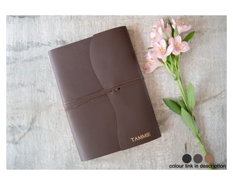 Könnte beinhalten: Ein braunes Lederjournal mit dem goldgeprägten Namen "TAMMIE" auf dem vorderen Umschlag. Das Journal ist geschlossen und mit einem braunen Lederband gebunden. Im Hintergrund befindet sich ein Strauß rosa Blumen.