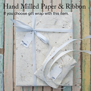 Puede incluir: Un regalo envuelto en papel blanco hecho a mano con una cinta blanca y dos etiquetas de regalo blancas. El texto "Hand Milled Paper & Ribbon" est&aacute; en la parte superior de la imagen. El texto "If you choose gift wrap with this item." est&aacute; debajo del primer texto.