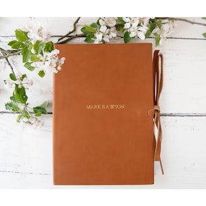 May include: A brown leather journal with a gold inscription that reads "Mark Rawson". The journal has a brown leather strap.