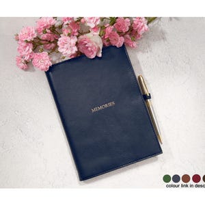May include: A navy blue journal with the word "MEMORIES" in gold lettering. A gold pen rests beside the journal, and pink flowers are arranged at the top. The journal is a classic design, perfect for recording thoughts and experiences.