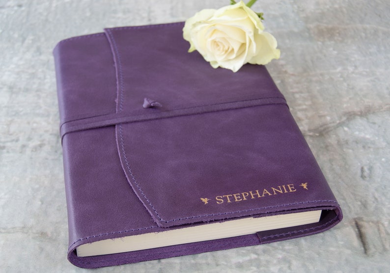 May include: A purple leather-bound journal with a gold-colored inscription that reads "STEPHANIE" and a white rose on top.
