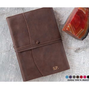 May include: A brown leather journal with a wrap-around strap closure and the initials S.P. embossed in the bottom right corner. Next to the journal is a polished, triangular-shaped red and orange agate stone with visible banding patterns.