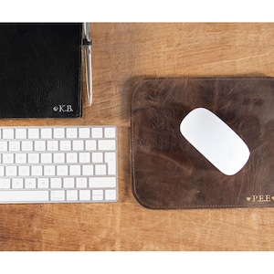 Puede incluir: Una alfombrilla de escritorio de cuero marrón con un ratón de ordenador blanco encima. La alfombrilla tiene el texto "♡ PEE ♡" en letras doradas.