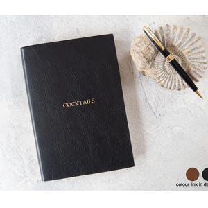 May include: A black leather-bound cocktail menu with the word "COCKTAILS" in gold lettering. A black and gold pen rests on a decorative stone. The menu and pen are on a light-colored surface.