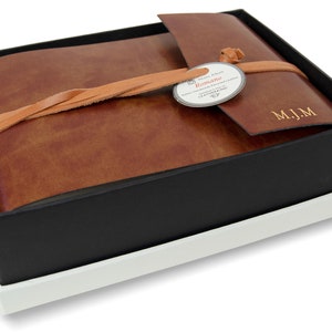 Puede incluir: Un &aacute;lbum de fotos encuadernado en cuero marr&oacute;n con cierre de solapa y una correa de cuero. El &aacute;lbum est&aacute; atado con un cord&oacute;n de cuero trenzado y tiene una etiqueta que dice "Romano" y "Hand Made in Italy". El &aacute;lbum est&aacute; monogramado con las iniciales "M.J.M."