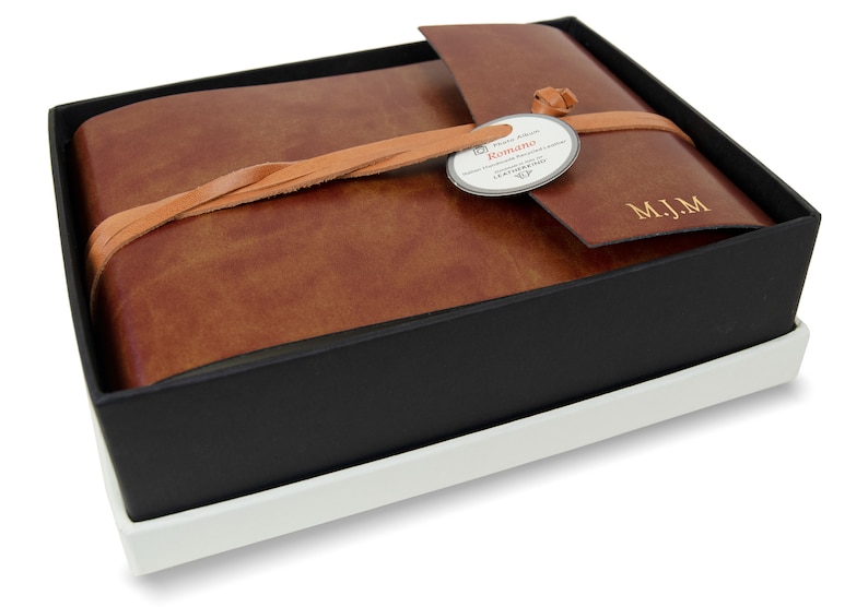 Puede incluir: Un &aacute;lbum de fotos encuadernado en cuero marr&oacute;n con cierre de solapa y una correa de cuero. El &aacute;lbum est&aacute; atado con un cord&oacute;n de cuero trenzado y tiene una etiqueta que dice "Romano" y "Hand Made in Italy". El &aacute;lbum est&aacute; monogramado con las iniciales "M.J.M."