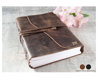May include: A dark brown leather journal with a wrap-around leather tie closure and visible side stitching. The front cover features the embossed initials JW in the bottom right corner. The journal contains white paper pages and has a rustic finish.