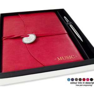 Puede incluir: Un juego de diario y bolígrafo de cuero rojo en una caja de regalo en blanco y negro. El diario está grabado con la palabra "MUSIC" en dorado. Se incluye un bolígrafo plateado. Una etiqueta está sujeta al diario con una cinta.