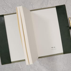 Puede incluir: Un cuaderno abierto con p&aacute;ginas blancas en blanco, encuadernado en una cubierta de cuero verde oscuro. Un bol&iacute;grafo dorado y plateado descansa en el lado derecho, sujeto por un lazo de cuero. El cuaderno est&aacute; sobre una superficie gris. El texto "Made in Italy" es visible.