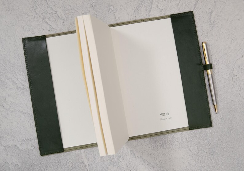 Puede incluir: Un cuaderno abierto con p&aacute;ginas blancas en blanco, encuadernado en una cubierta de cuero verde oscuro. Un bol&iacute;grafo dorado y plateado descansa en el lado derecho, sujeto por un lazo de cuero. El cuaderno est&aacute; sobre una superficie gris. El texto "Made in Italy" es visible.