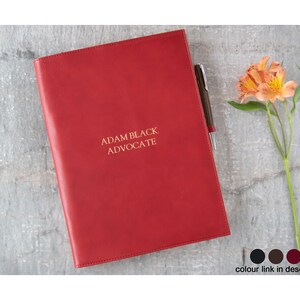 May include: Red leather folder with the text "ADAM BLACK ADVOCATE" embossed in gold. The folder is closed and lying flat on a gray surface.