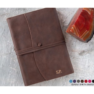 Könnte beinhalten: Ein braunes Lederjournal mit einem Riemenverschluss und den eingeprägten Initialen "S.P." auf der Vorderseite. Das Journal liegt auf einer strukturierten Oberfläche.