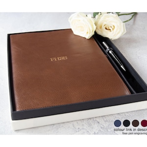 May include: A brown leather-textured notebook with the gold embossed letters FHSS on the cover, presented in a black and white gift box alongside a silver pen and two white roses. Five small colored circles are visible in the bottom right corner.