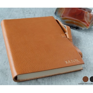 May include: A tan leather journal with the initials "M.S.S." embossed on the cover. The journal has a strap closure and a textured surface. A decorative stone is in the background.