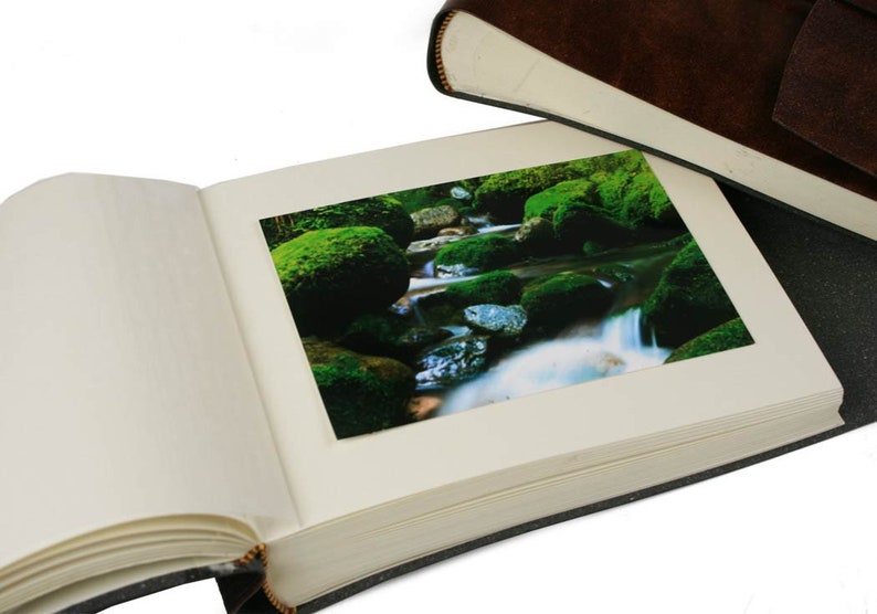 Puede incluir: Un &aacute;lbum de fotos de cuero marr&oacute;n con interior blanco y una foto de un arroyo cubierto de musgo con rocas insertadas en el &aacute;lbum.