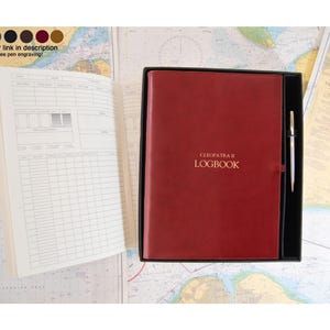 May include: A deep red leather-bound logbook titled CLEOPATRA II LOGBOOK in gold lettering, presented in a black box with a silver pen. An open logbook with white pages and printed grid tables is positioned to the left of the boxed set.