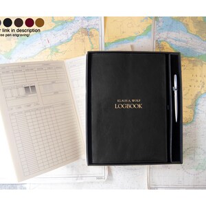 May include: A black logbook with the gold text KLAUS A. WOLF LOGBOOK on the cover, presented in a black box with a silver pen. An open logbook page with printed tables and columns is visible to the left, showing fields for date, time, and notes.