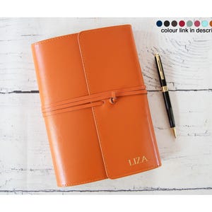 Peut inclure: Un journal en cuir orange avec une fermeture à lanière et le nom "LIZA" en relief doré. Un stylo noir et or est posé à côté du journal. Le journal est sur une surface en bois blanc.